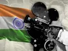 A photo montage of a colorful Indian flag waving on a white paper background, and a black & white photo of two Indian filmmakers peering through a large film movie camera.   